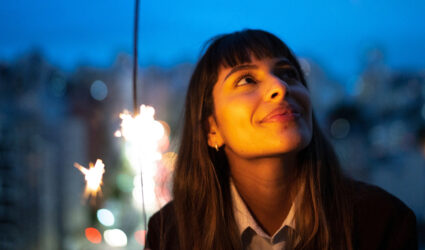 Smiling woman holding a sparkler with a blurred nighttime cityscape in the backdrop
