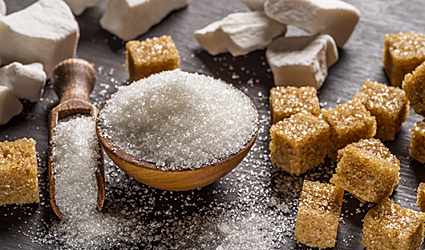 White sugar overflows from a small bowl and scoop onto a dark table surrounded by brown sugar cubes