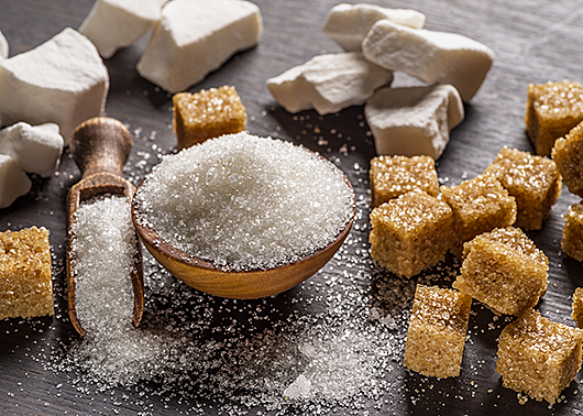 White sugar overflows from a small bowl and scoop onto a dark table surrounded by brown sugar cubes