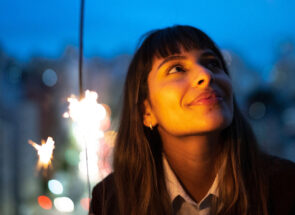 Smiling woman holding a sparkler with a blurred nighttime cityscape in the backdrop