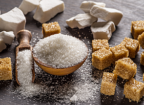 White sugar overflows from a small bowl and scoop onto a dark table surrounded by brown sugar cubes